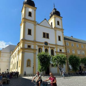 Two people riding bicycles in front of the historic Piarist Church in Trencin, Slovakia, on a sunny day during one day trip to Trencin