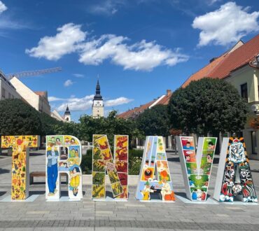 Colorful Trnava sign with artistic illustrations on a sunny day in the city center of Trnava, Slovakia, with historic buildings and a church in the background