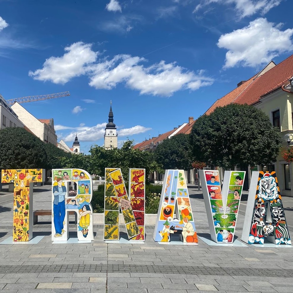 Colorful Trnava sign with artistic illustrations on a sunny day in the city center of Trnava, Slovakia, with historic buildings and a church in the background