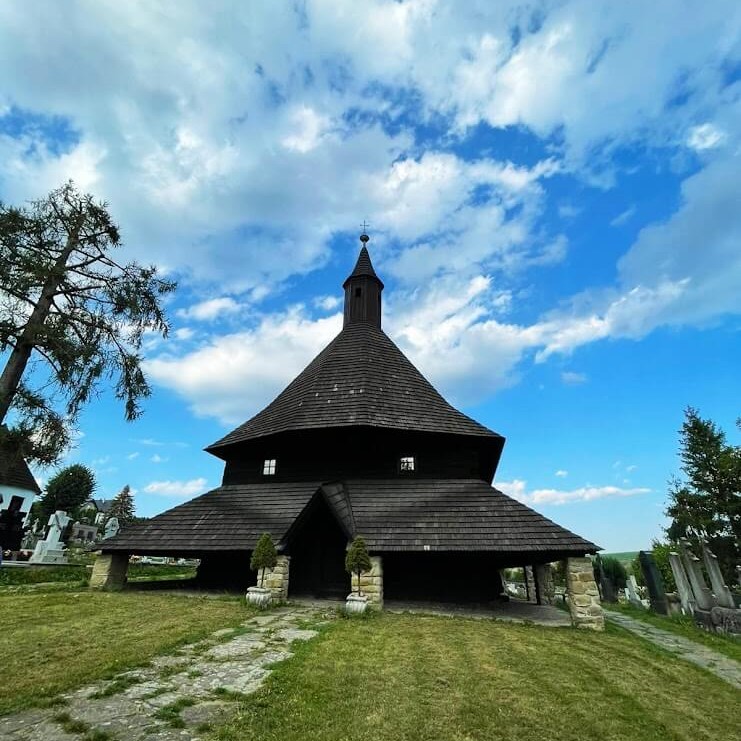 A historic wooden church with a unique conical roof stands against a bright blue sky in the Orava Region, Slovakia. The church's dark wooden structure contrasts beautifully with the lush green grass and the scattered clouds above, reflecting the traditional architecture and serene landscapes that define the Orava Region