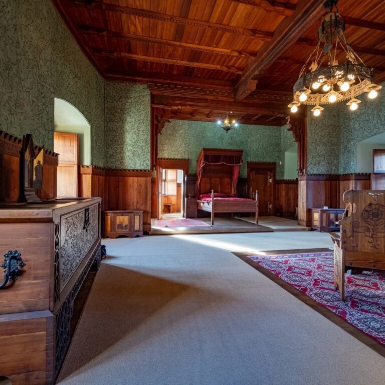 A beautifully preserved bedroom in Bojnice Castle, showcasing intricate woodwork and vintage decor. The centerpiece is a grand four-poster bed with deep red drapery, while the room's wooden walls, chandelier, and antique furnishings evoke a sense of regal history