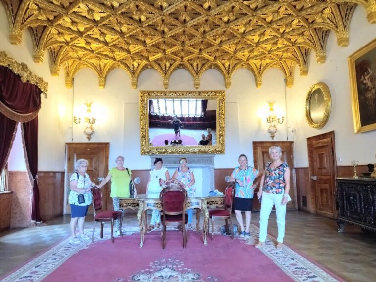 A group of visitors stands inside a luxurious room at Bojnice Castle, Slovakia. The room is adorned with a richly detailed golden ceiling and antique furnishings. A large ornate mirror reflects the room's elegance, while framed portraits and lavish decor enhance the historic ambiance