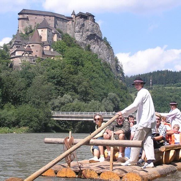 Tourists enjoy a traditional wooden raft ride on the Orava River, with the majestic Orava Castle towering in the background. The lush greenery surrounding the castle and the clear blue sky create a picturesque view, highlighting the natural and historical beauty of the Orava Region in Slovakia